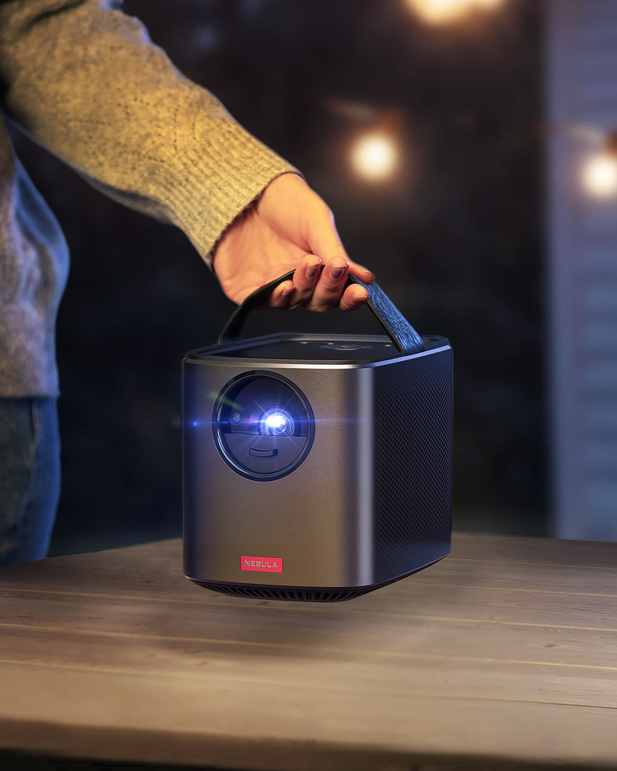 A hand uses the Nebula Mars II Pro handle to lift the portable projector off a wooden table.